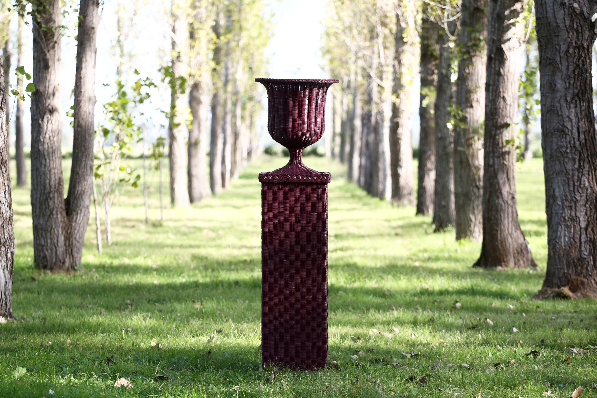 Handcrafted rattan urn-style vase with flowers, displayed among symmetrical rows of trees