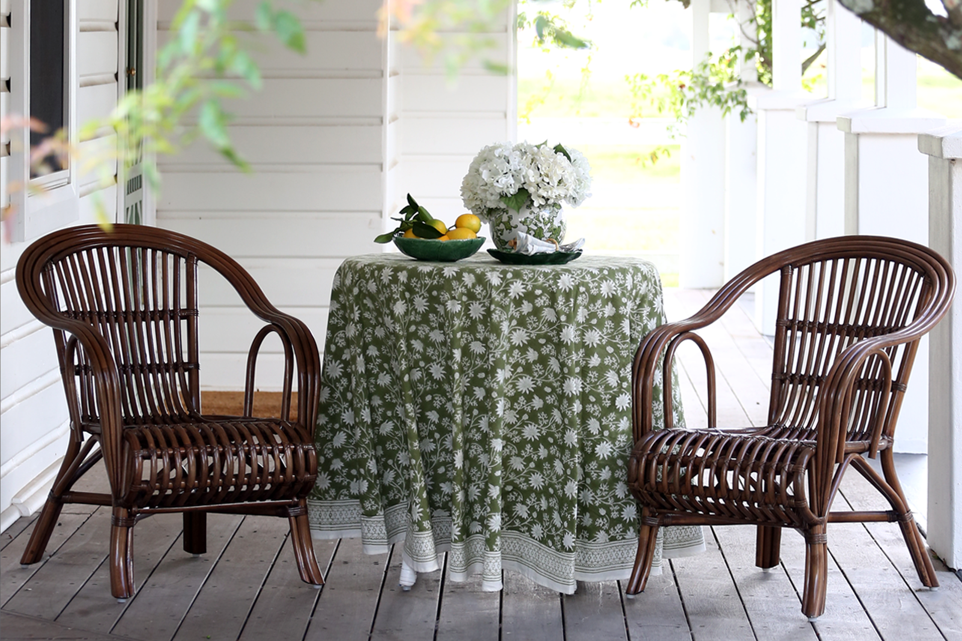 Handcrafted brown rattan chairs styled with a round table and floral tablecloth on a wooden veranda