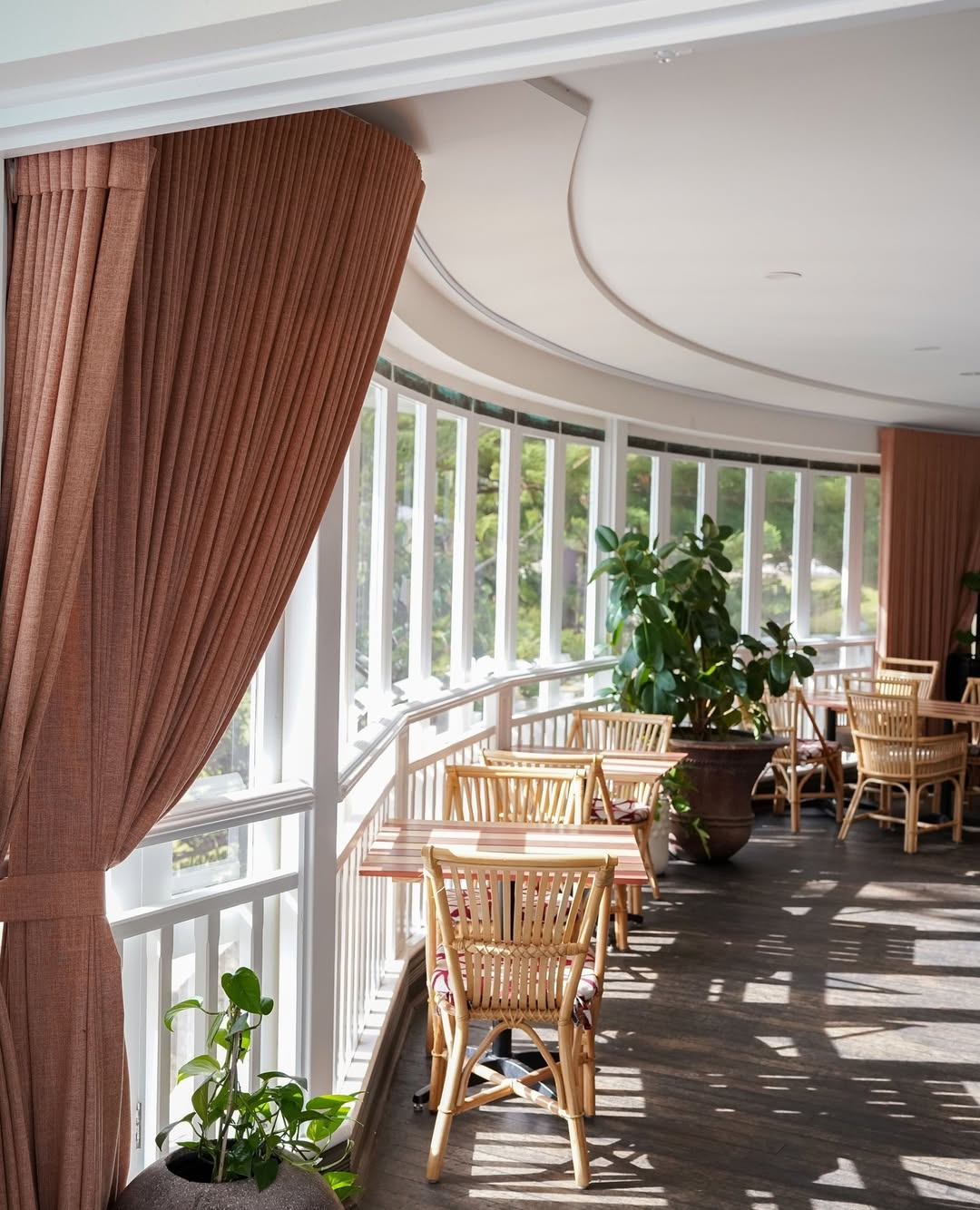 Sunlit indoor café space with light rattan dining chairs and wooden tables by curved floor-to-ceiling windows